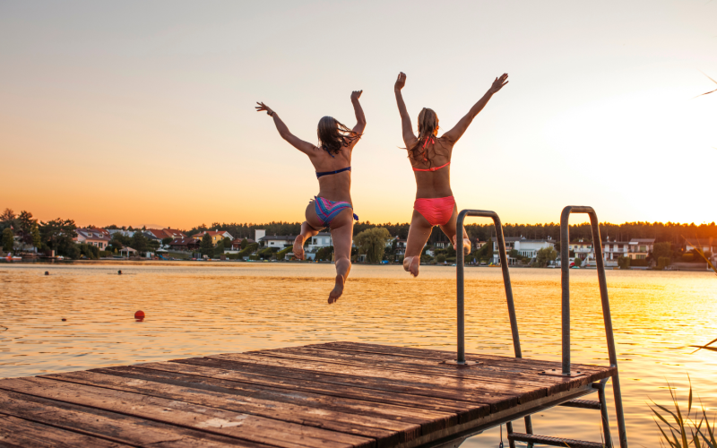To kvinner som hopper fra en brygge og ut i en sjø. Det er sommer og solnedgang.
