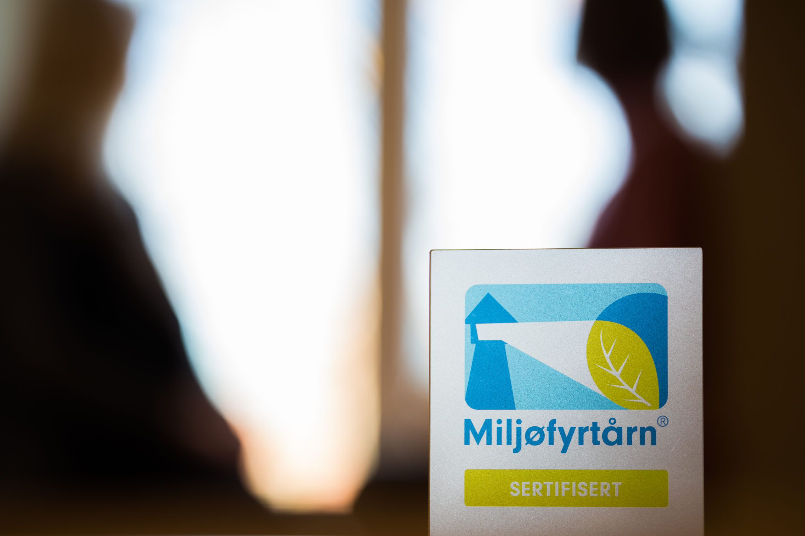 Miljøfyrtårn-plakett vises i fremgrunnen, med menneskeskikkelser i bakgrunn.