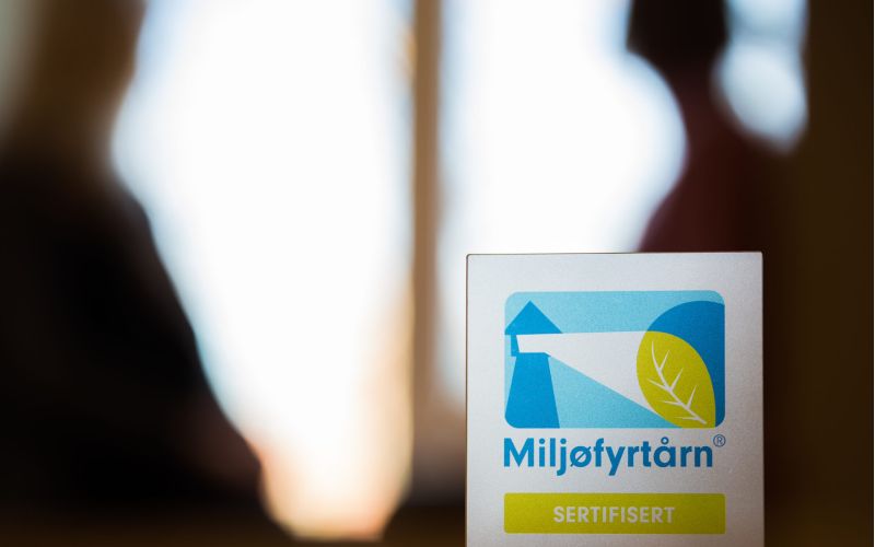 Miljøfyrtårn-plakett vises i fremgrunnen, med menneskeskikkelser i bakgrunn.