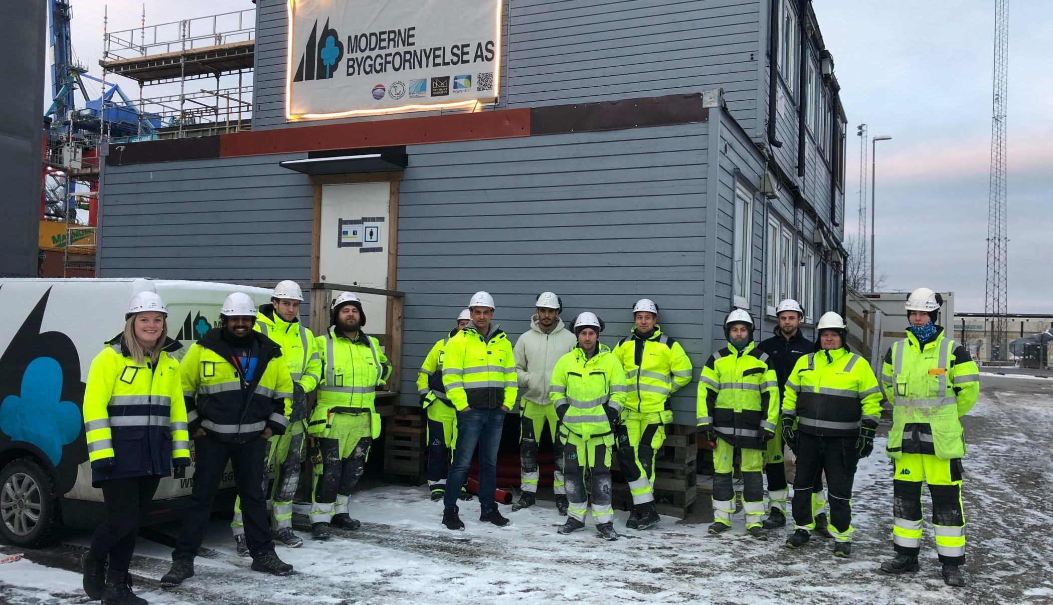 Byggeplass med anleggsarbeidere oppstilt foran varebil og brakker